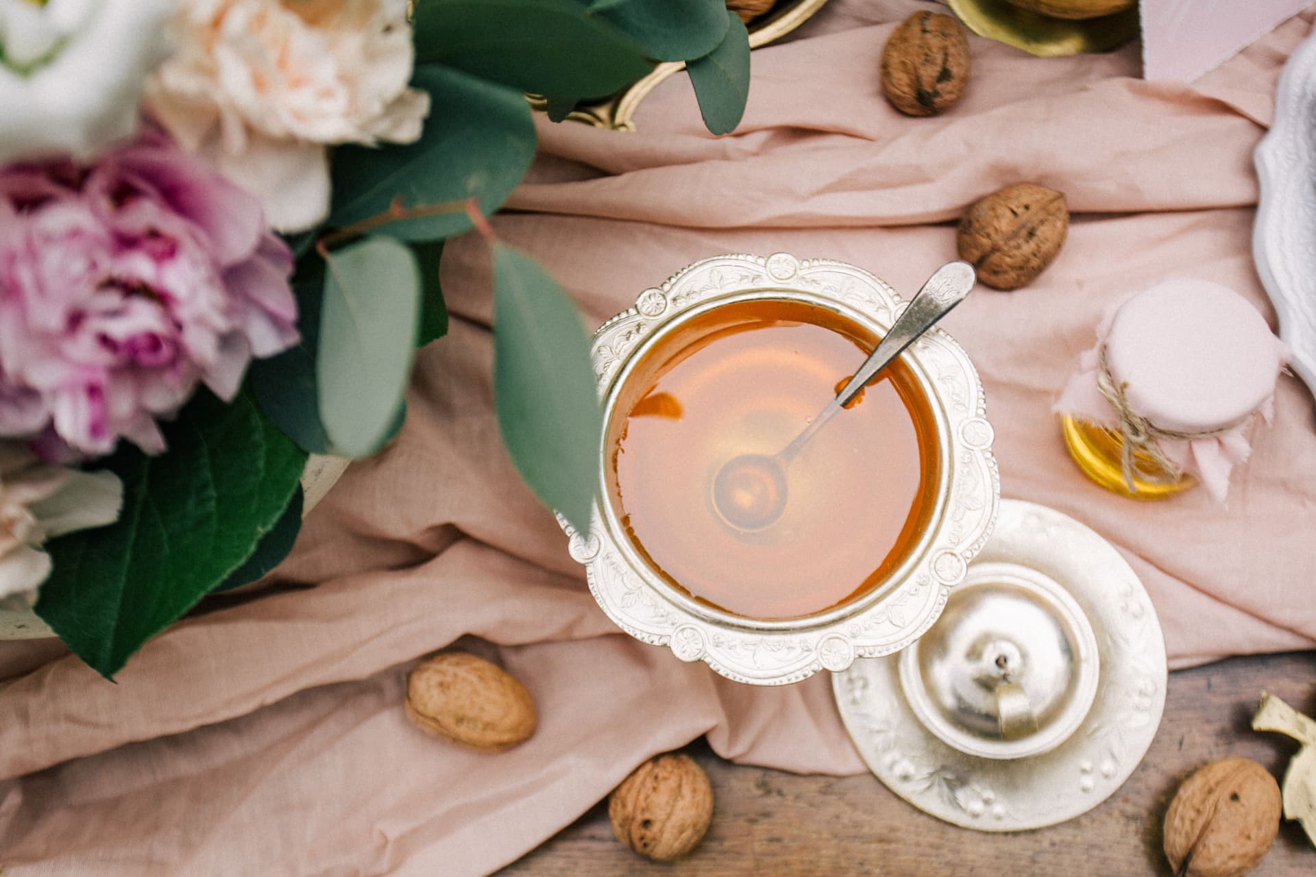 Amber honey in a silver bowl on blush linen with walnuts and peonies